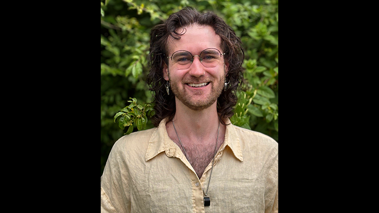 Jay Baker, in a yellow linen shirt, smiles at the camera while standing in front of a green leafy hedge.