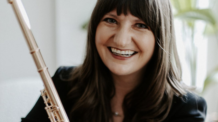 Fautist Cari Shipp, in black, sits in a white chair, holding her flute and smiles at the camera.